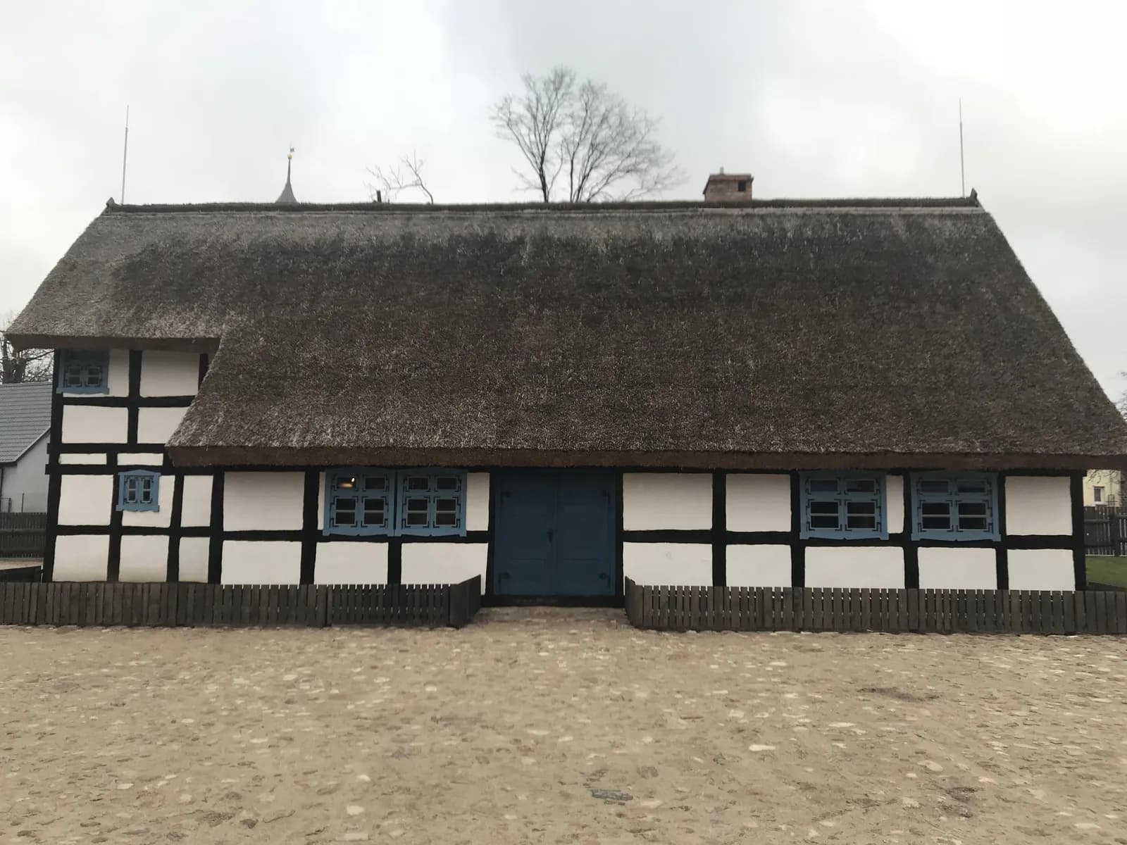 A traditional white house with exposed black timber framing, a thatched roof, and blue window frames and doors sits under an overcast sky.