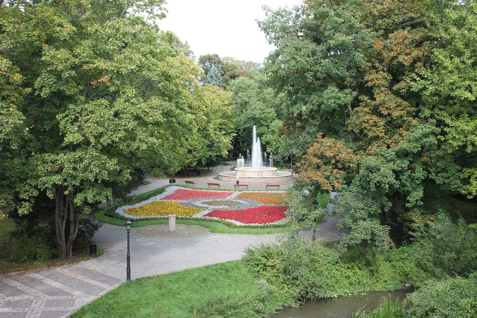 A park scene with a central fountain spraying water high into the air, surrounded by a large, colorful flower bed with geometric patterns of red, yellow, and other hues, all framed by lush green trees and winding pathways.
