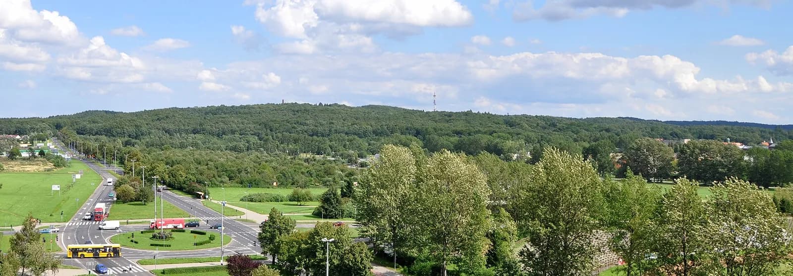 A panoramic view of a lush green landscape with a road, a roundabout with vehicles, and a dense forest in the background under a partly cloudy sky.